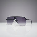 A pair of oversized shield-style sunglasses with a matte black frame and gradient gray lenses, displayed on a reflective white surface against a clean white background.