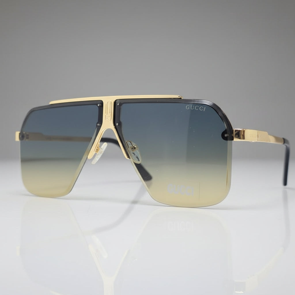 A pair of oversized shield-style sunglasses with gradient lenses that fade from blue-gray at the top to pale gold at the bottom, set in a gold-tone metal frame on a reflective white surface.