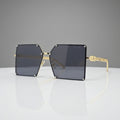 A pair of oversized square sunglasses with dark gray lenses and gold metal frames, displayed on a reflective white surface against a white background.