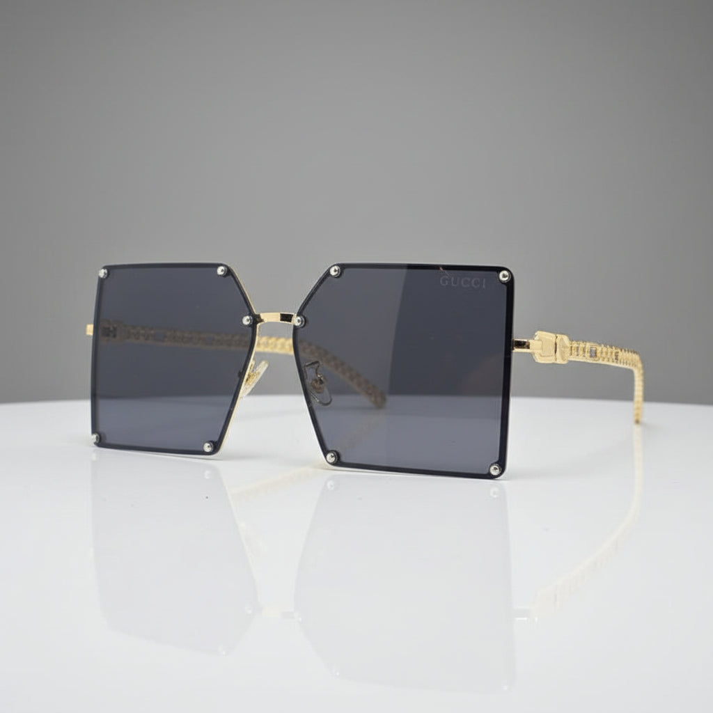 A pair of oversized square sunglasses with dark gray lenses and gold metal frames, displayed on a reflective white surface against a white background.