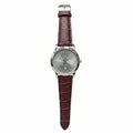 A wristwatch with a silver dial and a dark reddish-brown crocodile-embossed leather strap, held in a person's hand against a gray textured background.